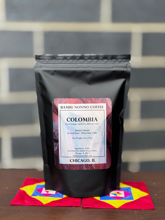 Black coffee bag with a label on a colorful mat against a gray tiled wall.