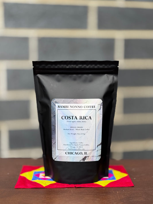 Black coffee bag labeled 'Costa Rica' from Bambi Nonno Coffee on a colorful mat with a gray brick wall background.