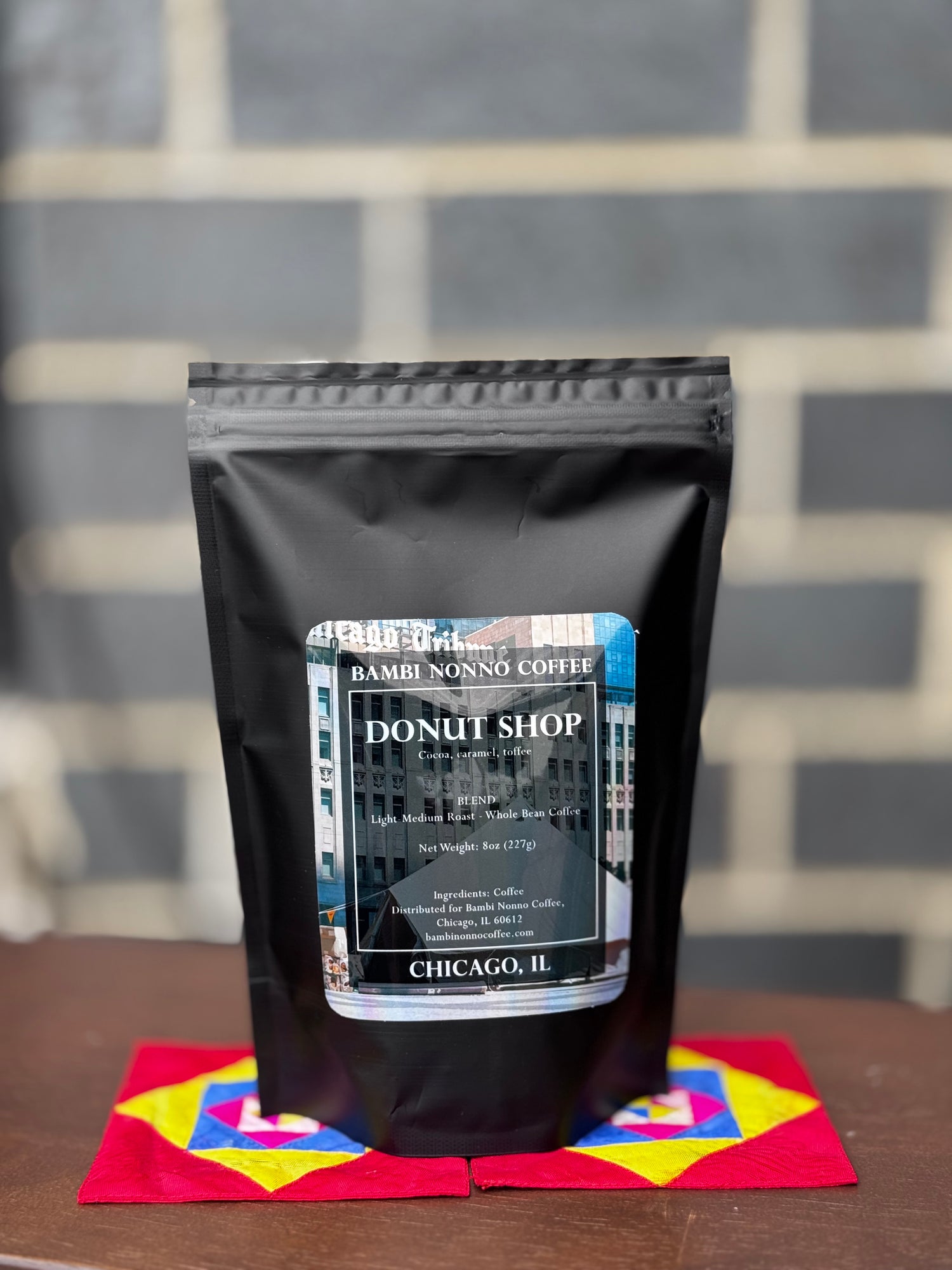 Black coffee bag labeled 'Donut Shop' on a colorful napkin with a gray brick wall background