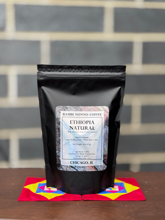 Black coffee bag labeled 'Bambi Nonno Coffee' with 'Ethiopia Natural' on a colorful mat against a brick wall.